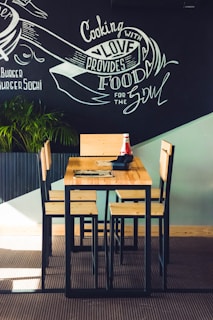 A wooden table with four matching chairs is set against a chalkboard wall featuring artistic writing that includes phrases like 'Cooking with love provides food for the soul.' A plant in the corner adds a touch of greenery, and there is a ketchup bottle and a napkin holder on the table.