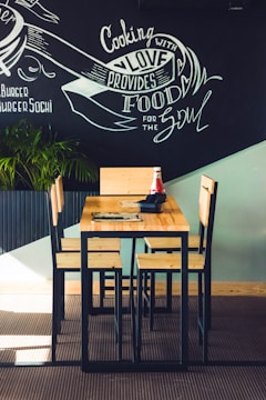 A wooden table with four matching chairs is set against a chalkboard wall featuring artistic writing that includes phrases like 'Cooking with love provides food for the soul.' A plant in the corner adds a touch of greenery, and there is a ketchup bottle and a napkin holder on the table.