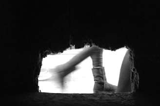 A black and white photo capturing a dynamic scene from a low perspective. The image shows a rough-edged hole with a view of blurry legs walking past, emphasizing movement. The scene has an abstract quality and a stark contrast between the dark foreground and bright background.