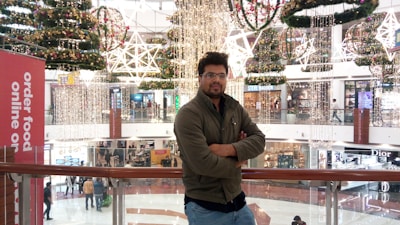 A person is standing on an upper floor of a shopping mall, leaning against a railing. The mall is elaborately decorated with hanging stars and festive wreaths with lights, suggesting a holiday season. Behind the person, multiple store signs are visible, including a large banner promoting online food ordering. Shoppers are visible on the lower level.