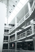 A multi-level modern interior with curved balconies and railings. The architecture features a striking combination of straight lines and rounded elements. The ceiling has large windows that allow natural light to brighten the space.