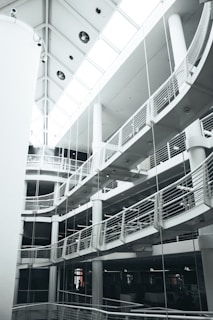 A multi-level modern interior with curved balconies and railings. The architecture features a striking combination of straight lines and rounded elements. The ceiling has large windows that allow natural light to brighten the space.