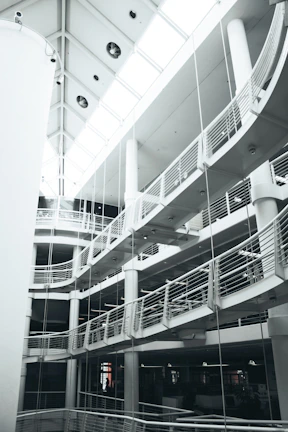 A multi-level modern interior with curved balconies and railings. The architecture features a striking combination of straight lines and rounded elements. The ceiling has large windows that allow natural light to brighten the space.