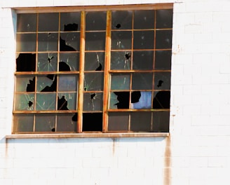 A window with multiple panes of glass, many of which are broken, is set within a white brick wall. The frames are wooden and show signs of weathering.