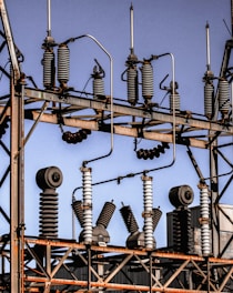 A series of industrial electrical insulators and transformers positioned on a grid-like metal structure. The arrangement is orderly, with cylindrical and spiral shapes dominating the composition. The sky in the background is clear and blue.