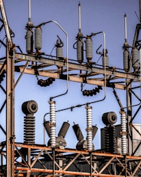 A series of industrial electrical insulators and transformers positioned on a grid-like metal structure. The arrangement is orderly, with cylindrical and spiral shapes dominating the composition. The sky in the background is clear and blue.
