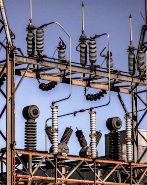 A series of industrial electrical insulators and transformers positioned on a grid-like metal structure. The arrangement is orderly, with cylindrical and spiral shapes dominating the composition. The sky in the background is clear and blue.