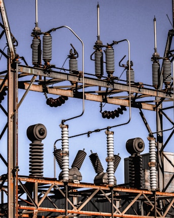 A series of industrial electrical insulators and transformers positioned on a grid-like metal structure. The arrangement is orderly, with cylindrical and spiral shapes dominating the composition. The sky in the background is clear and blue.