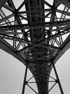 A black and white photo taken from beneath a large steel bridge, showcasing a complex network of intersecting beams and trusses. The intricate pattern of the metalwork is highlighted against the bright sky, emphasizing the engineering and architectural design.