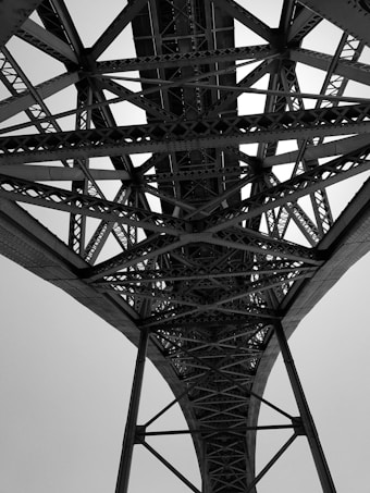 A black and white photo taken from beneath a large steel bridge, showcasing a complex network of intersecting beams and trusses. The intricate pattern of the metalwork is highlighted against the bright sky, emphasizing the engineering and architectural design.