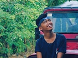 A person with a headband and a joyful expression is looking upwards while sitting outdoors. Behind him, there is a red vehicle partially covered with rain droplets on the window. The background is filled with lush green foliage, adding to the vibrant and natural setting.