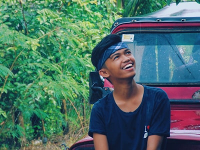 A person with a headband and a joyful expression is looking upwards while sitting outdoors. Behind him, there is a red vehicle partially covered with rain droplets on the window. The background is filled with lush green foliage, adding to the vibrant and natural setting.
