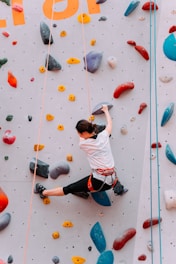 A person is climbing an indoor rock climbing wall covered with various colored holds. They are wearing a climbing harness and shoes, and are using both hands and feet to ascend the wall.