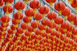 A large collection of red Chinese lanterns hanging in rows, adorned with yellow tassels. Each lantern has black Chinese characters and designs on the surface. The background features a blue-tinted building with windows, creating a festive ambiance.