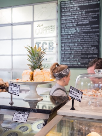 A dessert display in a deli featuring a plate of pi&ntilde;a colada cake topped with a small pineapple, with a banana split cake under a glass dome. There are signs indicating the cake names and prices. A person in the background is wearing a headband.