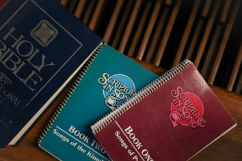 A group of religious books resting on a wooden surface. The blue book is a Holy Bible, while two spiral-bound books titled 'Scripture in Song' in teal and red are nearby, labeled as Book Two and Book One, respectively. The books have a vintage appearance.