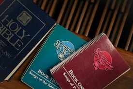 A group of religious books resting on a wooden surface. The blue book is a Holy Bible, while two spiral-bound books titled 'Scripture in Song' in teal and red are nearby, labeled as Book Two and Book One, respectively. The books have a vintage appearance.