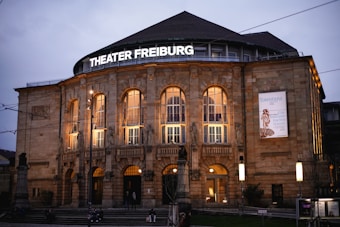 The image captures the exterior of a grand theater building during the evening. The facade is illuminated with warm lights highlighting architectural details such as columns and statues. Large arched windows line the front, and there is a prominent sign displaying 'Theater Freiburg.' A poster is visible on one side of the building advertising a performance. People are gathered near the entrance and on the steps.
