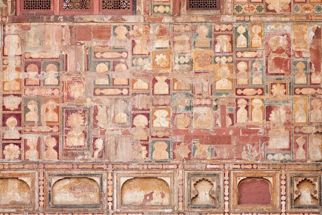 The image features a richly decorated, ornate wall showcasing intricate patterns and faded frescoes. The design consists of repetitive geometric shapes and floral motifs in a variety of earthy tones. The top section includes small, lattice-covered windows.