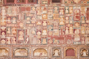 The image features a richly decorated, ornate wall showcasing intricate patterns and faded frescoes. The design consists of repetitive geometric shapes and floral motifs in a variety of earthy tones. The top section includes small, lattice-covered windows.