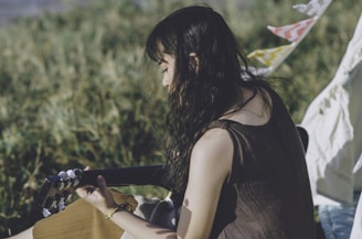 A person with long, wavy dark hair is playing an acoustic guitar outdoors. The background consists of lush greenery and blurred plants, suggesting a serene, natural setting. Colorful flags or fabric streamers are visible, adding a touch of festivity.