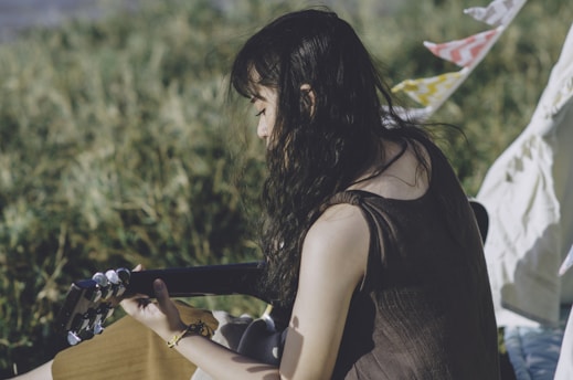 A person with long, wavy dark hair is playing an acoustic guitar outdoors. The background consists of lush greenery and blurred plants, suggesting a serene, natural setting. Colorful flags or fabric streamers are visible, adding a touch of festivity.