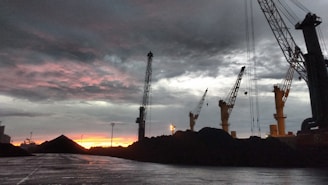 Cranes towering over heaps of coal with a dramatic, cloudy sky and a vibrant reddish-orange sunset in the background. The scene is industrial with a focus on the machinery against the natural backdrop.