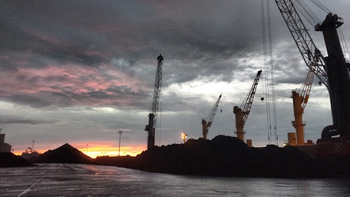 Cranes towering over heaps of coal with a dramatic, cloudy sky and a vibrant reddish-orange sunset in the background. The scene is industrial with a focus on the machinery against the natural backdrop.