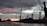 Cranes towering over heaps of coal with a dramatic, cloudy sky and a vibrant reddish-orange sunset in the background. The scene is industrial with a focus on the machinery against the natural backdrop.