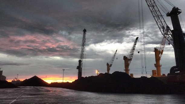 Cranes towering over heaps of coal with a dramatic, cloudy sky and a vibrant reddish-orange sunset in the background. The scene is industrial with a focus on the machinery against the natural backdrop.