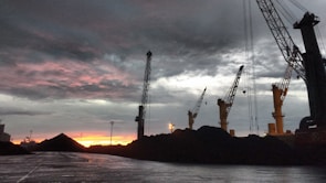 Cranes towering over heaps of coal with a dramatic, cloudy sky and a vibrant reddish-orange sunset in the background. The scene is industrial with a focus on the machinery against the natural backdrop.