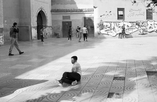 Children and adults engage in various activities in a plaza with intricate tile patterns. A child sits on the ground in the foreground, while others play near a wall featuring graffiti and Arabic script.