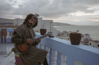 A man with dreadlocks and a beard is playing the guitar on a rooftop terrace. The setting is serene with several potted plants placed on the blue railing. The background reveals a coastal cityscape with buildings and a body of water under a cloudy sky.