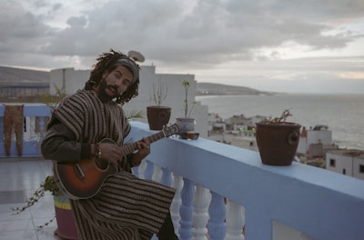 A man with dreadlocks and a beard is playing the guitar on a rooftop terrace. The setting is serene with several potted plants placed on the blue railing. The background reveals a coastal cityscape with buildings and a body of water under a cloudy sky.