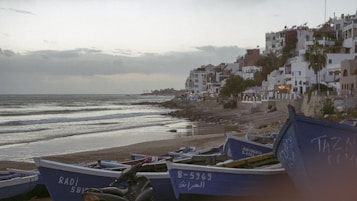 The shoreline features a row of blue fishing boats resting on the sand, with a tranquil sea in the background. To the right, a series of white and pastel-colored buildings are perched on a hillside, suggesting a coastal village or town. The sky is overcast, giving off a muted, serene vibe.