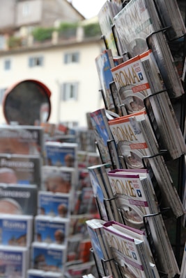 A display rack with various tourist magazines, featuring covers related to Rome. The magazines are neatly arranged and prominently feature images and text. In the background, there's a blurred view of a building and more magazines.
