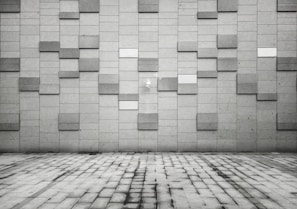 A geometric pattern wall composed of rectangular tiles in various shades of gray. The foreground features a pavement made of rectangular slabs, mirroring the design of the wall. The overall aesthetic is modern and minimalist, with a subtle play of light and shadow across the surfaces.