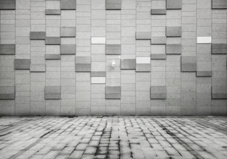 A geometric pattern wall composed of rectangular tiles in various shades of gray. The foreground features a pavement made of rectangular slabs, mirroring the design of the wall. The overall aesthetic is modern and minimalist, with a subtle play of light and shadow across the surfaces.