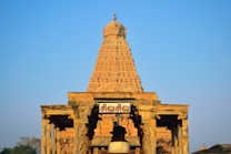 A large, intricately carved temple structure with a detailed tower and architectural elements typical of ancient Indian design. The stone facade is illuminated by a soft golden light against a clear blue sky. A sign with text in a local script is prominently displayed.