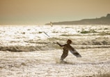 A person engaged in kite surfing is captured amidst ocean waves under a golden sky. The warm sunlight reflects off the water, creating a sparkling effect. The silhouette of the surfer is pronounced against the glistening sea and expansive horizon.