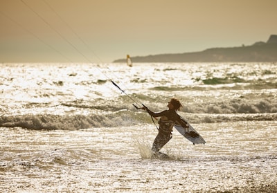 A person engaged in kite surfing is captured amidst ocean waves under a golden sky. The warm sunlight reflects off the water, creating a sparkling effect. The silhouette of the surfer is pronounced against the glistening sea and expansive horizon.