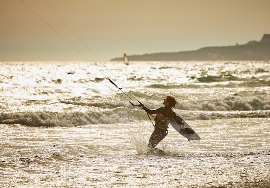 A person engaged in kite surfing is captured amidst ocean waves under a golden sky. The warm sunlight reflects off the water, creating a sparkling effect. The silhouette of the surfer is pronounced against the glistening sea and expansive horizon.