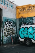 A graffiti-covered wall features various colorful tags and patterns. The prominent colors include teal, yellow, and black. A white van parked nearby displays its own graffiti, matching the style of the surrounding walls. The walls belong to a vehicle repair shop, indicated by signs advertising replacements for automotive parts.