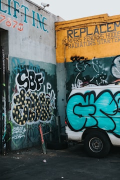 A graffiti-covered wall features various colorful tags and patterns. The prominent colors include teal, yellow, and black. A white van parked nearby displays its own graffiti, matching the style of the surrounding walls. The walls belong to a vehicle repair shop, indicated by signs advertising replacements for automotive parts.