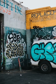 A graffiti-covered wall features various colorful tags and patterns. The prominent colors include teal, yellow, and black. A white van parked nearby displays its own graffiti, matching the style of the surrounding walls. The walls belong to a vehicle repair shop, indicated by signs advertising replacements for automotive parts.