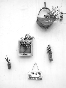 A minimalist wall display featuring various decorative items. A small basket labeled 'ALLSEASONS' holds some flowers, while a wooden frame with the word 'Home' contains a bouquet of flowers. To the left, a simple piece of wood holds a small artificial cactus. Below, a rustic sign with Chinese characters adds a touch of cultural detail. Overall, the composition is artistic and clean.