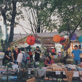 A vibrant market scene featuring a stall decorated with colorful umbrellas and flowers. People are interacting and there are various goods like food items displayed. The atmosphere is lively and festive with greenery and trees surrounding the area.