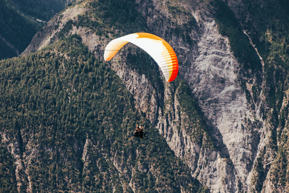The image depicts a paraglider suspended in mid-air, soaring above a mountainous landscape. The paraglider is painted in vibrant shades of orange and white, with an orange parachute trailing behind it. Below, the mountain range stretches across the scene, its rocky peaks contrasting against the clear blue sky. The perspective of the image is from below, looking up at the paraglider and the mountains, creating a sense of awe and adventure.