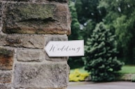 A white arrow-shaped sign with the word 'Wedding' written in elegant, cursive script is attached to a rustic stone wall. The background features lush greenery, including a large tree and various plants.