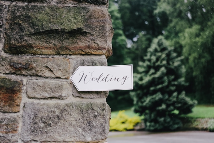 A white arrow-shaped sign with the word 'Wedding' written in elegant, cursive script is attached to a rustic stone wall. The background features lush greenery, including a large tree and various plants.